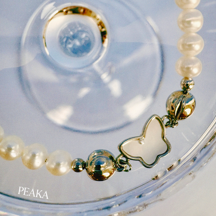 Pearl bracelet with a butterfly charm on a clear background, branded 'PEAKA'.